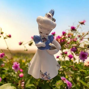 May include: A vintage ceramic figurine of a woman in a white dress with blue accents and floral details. The figurine is holding a basket and wearing a hat. The background is a blurred image of flowers and a blue sky.