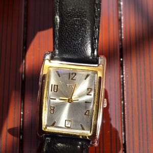 May include: A rectangular Guess watch with a silver face, gold-colored bezel, and black leather band. The watch face displays the numbers 2, 4, 8, 10, and 12, along with the date. The watch is a classic accessory.