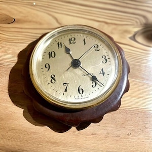 May include: A round quartz clock with a gold-colored face and black hands and numbers. The clock is set in a dark brown, scalloped wooden frame. The text "QUARTZ" and "MADE IN FRANCE" are visible on the clock face.