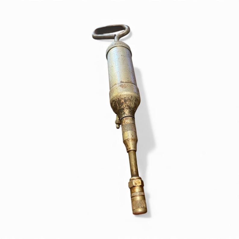 Pode incluir: Uma pistola de graxa antiga com corpo prateado e bico de lat&atilde;o. A al&ccedil;a tem formato de la&ccedil;o. A pistola mostra sinais de desgaste, com alguma ferrugem e descolora&ccedil;&atilde;o. &Eacute; projetada para dispensar graxa.