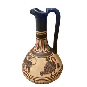 May include: A ceramic jug with a black handle and spout, featuring a beige body adorned with ancient Greek-style illustrations in brown and red. The jug has a decorative pattern around the neck and base, with a lion and bird design.