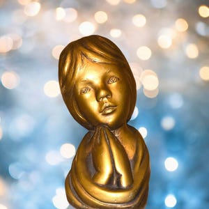 May include: A golden-colored statue of a child with hands clasped in prayer. The sculpture has detailed facial features and is set against a blurred background of blue and gold lights. The statue is a religious or decorative item.