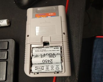 May include: A white and gray Game Boy Color handheld gaming console with a clear plastic battery compartment. The compartment contains three AA batteries labeled "LADDA 2450", "IKEA 23076", and "23078".