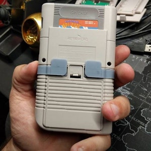 Könnte beinhalten: Eine graue Game Boy Color Handheld-Spielkonsole mit einer grauen Kassette, die eingesetzt ist. Die Kassette trägt den Text "CAPCOM" und "STREET FIGHTER II". Die Konsole hat ein graues Kunststoffgehäuse mit einer strukturierten Oberfläche. Die Konsole ist mit "RETROFLAG" und "GP CASE" beschriftet.