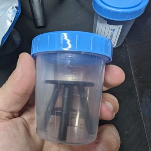 May include: A clear plastic container with a blue lid and a black tripod stand inside. The container has markings for measuring volume in milliliters.