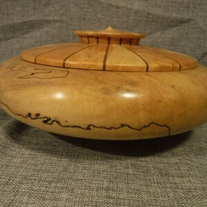 Könnte beinhalten: Eine Holzschale mit Deckel. Die Schale ist aus hellem Holz mit dunkelbraunen Streifen. Der Deckel hat ein gestreiftes Muster.