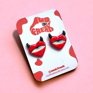 Può includere: Un paio di orecchini a forma di cuore rosso con un occhio bianco e corna di diavolo nere. Gli orecchini sono su una carta bianca con il testo "mud freak" e le maniglie dei social media.