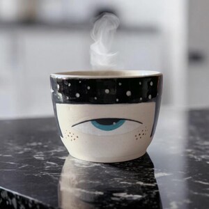 Peut inclure: Une tasse en céramique blanche avec un motif noir et blanc. La tasse a un seul œil bleu peint sur le devant. De la vapeur s'élève de la tasse.