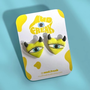 May include: Yellow heart-shaped earrings with black horns and an eye design, presented on a white card. The card features the text "MUD FREAK" and the brand's social media details and website address.