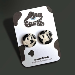 Puede incluir: Un par de pendientes con carita sonriente de estampado de vaca en blanco y negro. Los pendientes están en una tarjeta blanca con el texto "mud.freak" y una dirección de sitio web.