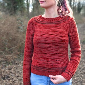 Op de afbeelding: Een vrouw draagt een rode gehaakte trui met een ronde hals en lange mouwen. De afbeelding bevat een tekstoverlay met de tekst "Oletta Sweater" en "A PDF Crochet Pattern in English and in Dutch".