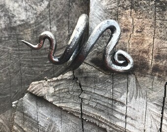 May include: A silver metal ring shaped like a snake with a curled tail. The ring is on a wooden surface.