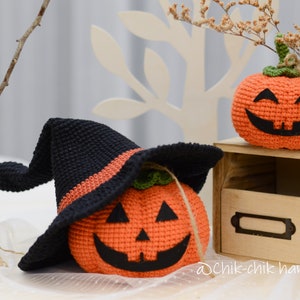 May include: Two crocheted pumpkins, one wearing a black witch hat with an orange band. The pumpkins have orange bodies with black facial features and green stems. The pumpkins are sitting on a wooden shelf with a label that says "@Chik-chik handmade".