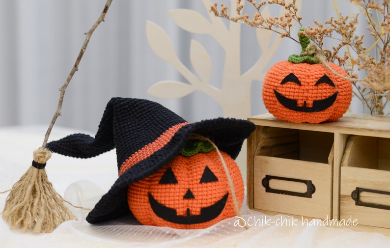 May include: Two crocheted pumpkins, one wearing a black witch hat with an orange band. The pumpkins have orange bodies with black facial features and green stems. The pumpkins are sitting on a wooden shelf with a label that says "@Chik-chik handmade".