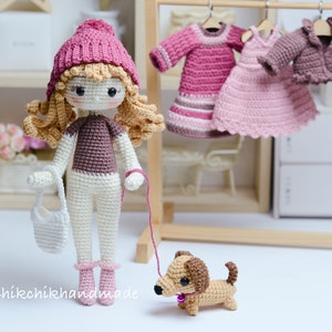 May include: A crocheted doll wearing a pink hat and brown sweater, holding a white bag and a red lead attached to a brown and tan crocheted dachshund. The doll is standing on a white surface in front of a wooden shelf with a pink and white crocheted dress hanging on a hanger.