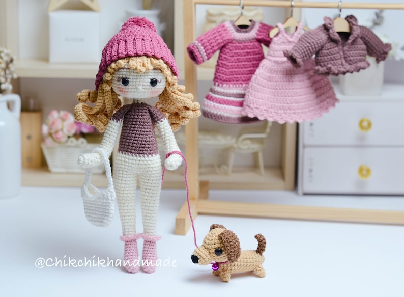 May include: A crocheted doll wearing a pink hat and brown sweater, holding a white bag and a red lead attached to a brown and tan crocheted dachshund. The doll is standing on a white surface in front of a wooden shelf with a pink and white crocheted dress hanging on a hanger.
