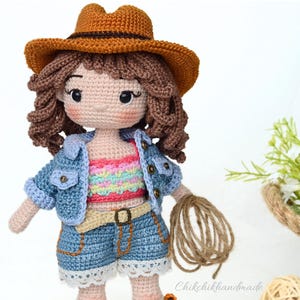 K&ouml;nnte beinhalten: Eine geh&auml;kelte Puppe, die eine Jeansjacke, ein regenbogenfarbenes gestreiftes Oberteil, Jeans-Shorts und einen braunen Cowboyhut tr&auml;gt. Die Puppe h&auml;lt eine Kn&auml;uel Schnur.