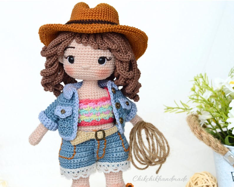 K&ouml;nnte beinhalten: Eine geh&auml;kelte Puppe, die eine Jeansjacke, ein regenbogenfarbenes gestreiftes Oberteil, Jeans-Shorts und einen braunen Cowboyhut tr&auml;gt. Die Puppe h&auml;lt eine Kn&auml;uel Schnur.