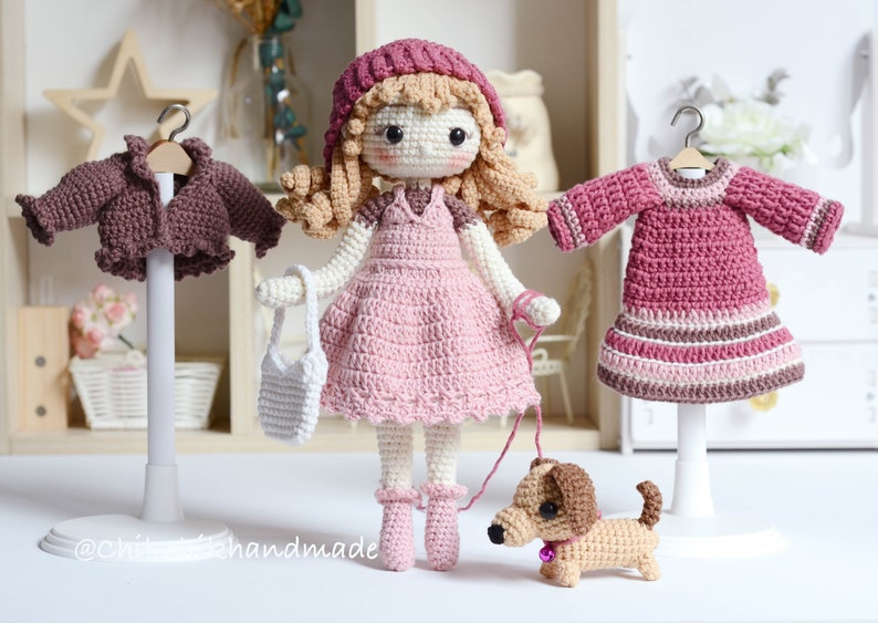May include: Crocheted doll wearing a pink dress, brown hat, and white handbag, holding a lead attached to a brown crocheted dachshund. The doll is standing on a white surface with two other crocheted garments on stands behind it.