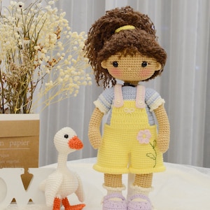 May include: A crocheted doll with brown hair in a ponytail wearing a blue and white striped shirt and yellow overalls with a pink flower. The doll is standing next to a white crocheted goose with orange feet.