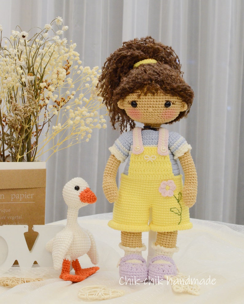May include: A crocheted doll with brown hair in a ponytail wearing a blue and white striped shirt and yellow overalls with a pink flower. The doll is standing next to a white crocheted goose with orange feet.