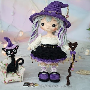 May include: A crocheted doll with long light gray hair wearing a purple witch hat, a white off-shoulder top, and a black skirt. The doll holds a brown heart-topped staff. Beside it sits a small black cat in a floral armchair with a purple hat.