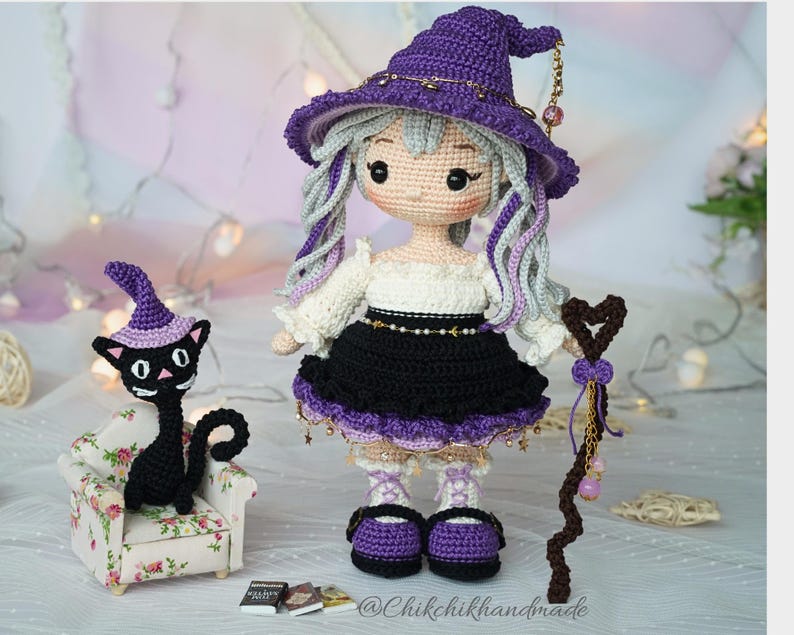 May include: A crocheted doll with long light gray hair wearing a purple witch hat, a white off-shoulder top, and a black skirt. The doll holds a brown heart-topped staff. Beside it sits a small black cat in a floral armchair with a purple hat.