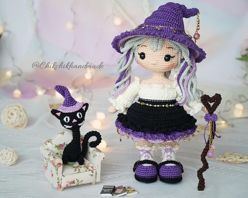 May include: A crochet doll wearing a purple witch hat, white blouse, and black skirt, holding a brown heart-topped staff. Beside the doll is a black crochet cat in a floral armchair, wearing a purple hat, with three miniature books on the floor.