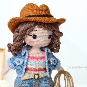 K&ouml;nnte beinhalten: H&auml;kelpuppe mit braunem Cowboyhut, blauer Jeansjacke und einem bunten gestreiften Oberteil. Die Puppe hat braunes Haar und h&auml;lt ein Seil.
