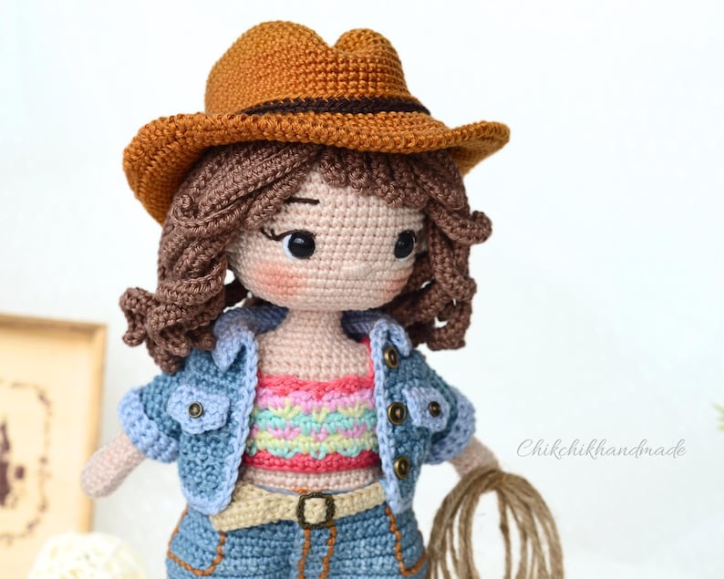 K&ouml;nnte beinhalten: H&auml;kelpuppe mit braunem Cowboyhut, blauer Jeansjacke und einem bunten gestreiften Oberteil. Die Puppe hat braunes Haar und h&auml;lt ein Seil.