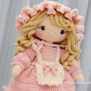 Pode incluir: Uma boneca de croch&ecirc; com cabelo loiro, vestindo um vestido rosa e um avental branco com um la&ccedil;o rosa. A boneca est&aacute; usando um chap&eacute;u de croch&ecirc; rosa com acabamento branco. A boneca &eacute; feita &agrave; m&atilde;o e tem uma express&atilde;o doce.