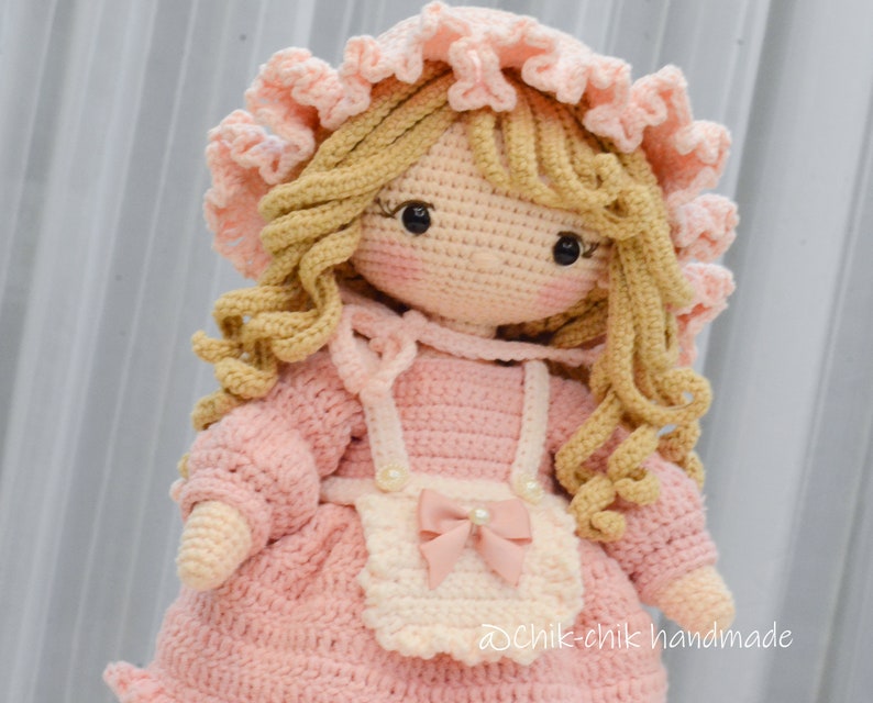 Pode incluir: Uma boneca de croch&ecirc; com cabelo loiro, vestindo um vestido rosa e um avental branco com um la&ccedil;o rosa. A boneca est&aacute; usando um chap&eacute;u de croch&ecirc; rosa com acabamento branco. A boneca &eacute; feita &agrave; m&atilde;o e tem uma express&atilde;o doce.