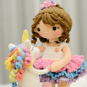 May include: A crocheted doll wearing a pink and blue dress with a white unicorn toy. The doll has brown hair and a flower crown. The unicorn has a rainbow mane and tail.