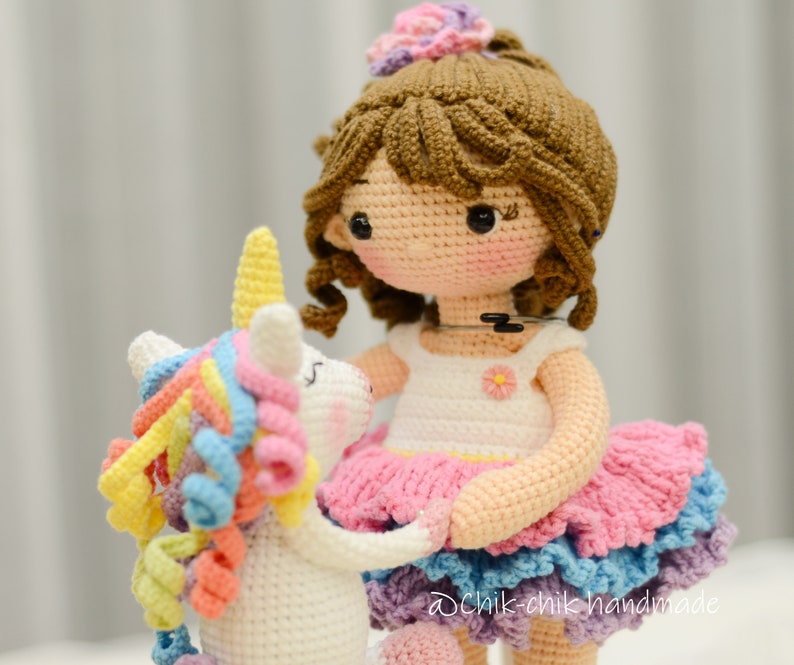 May include: A crocheted doll wearing a pink and blue dress with a white unicorn toy. The doll has brown hair and a flower crown. The unicorn has a rainbow mane and tail.