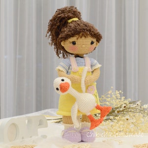 May include: A crocheted doll with brown hair wearing a yellow dress and holding a white crocheted goose. The doll is standing on a white surface with a white "LOVE" sign in the foreground.