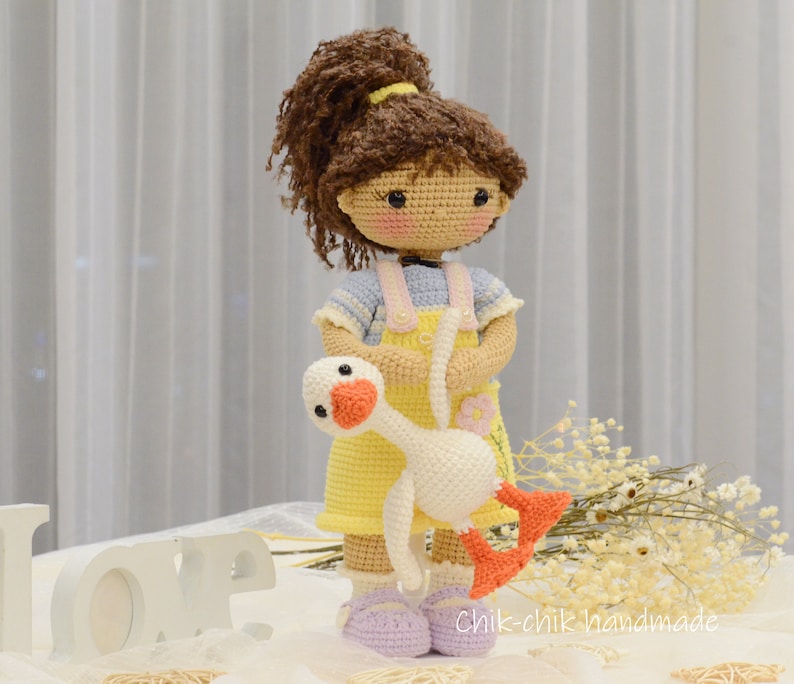 May include: A crocheted doll with brown hair wearing a yellow dress and holding a white crocheted goose. The doll is standing on a white surface with a white "LOVE" sign in the foreground.
