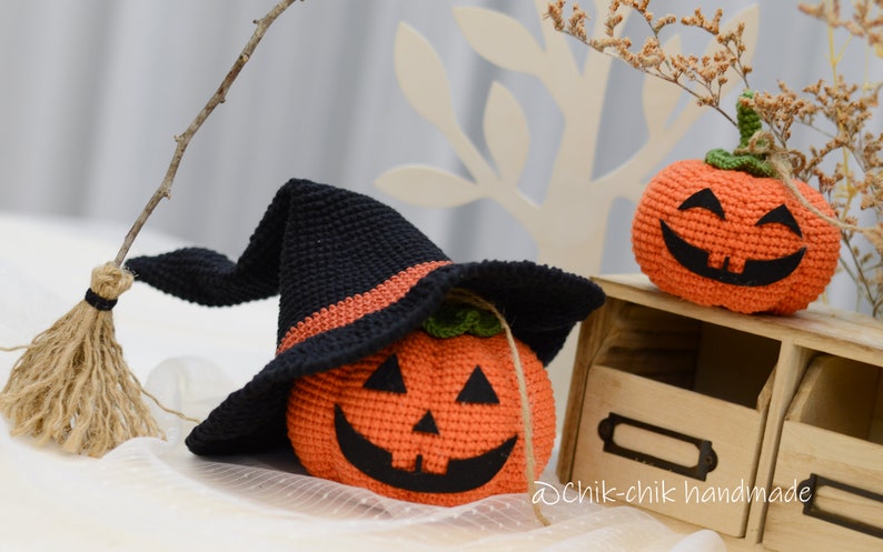 May include: Two crocheted pumpkins, one wearing a black witch hat with an orange stripe. The pumpkins have smiling faces with black eyes and mouths.  The pumpkins are sitting on a wooden shelf with a label that says "Dchik-chik handmade".