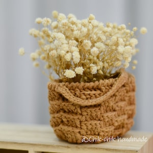Pode incluir: Uma pequena cesta de croch&ecirc; marrom cheia de flores brancas secas. A cesta tem uma textura tecida e um design simples. As flores s&atilde;o delicadas e adicionam um toque de beleza natural &agrave; disposi&ccedil;&atilde;o.