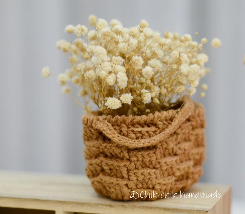 Pode incluir: Uma pequena cesta de croch&ecirc; marrom cheia de flores brancas secas. A cesta tem uma textura tecida e um design simples. As flores s&atilde;o delicadas e adicionam um toque de beleza natural &agrave; disposi&ccedil;&atilde;o.