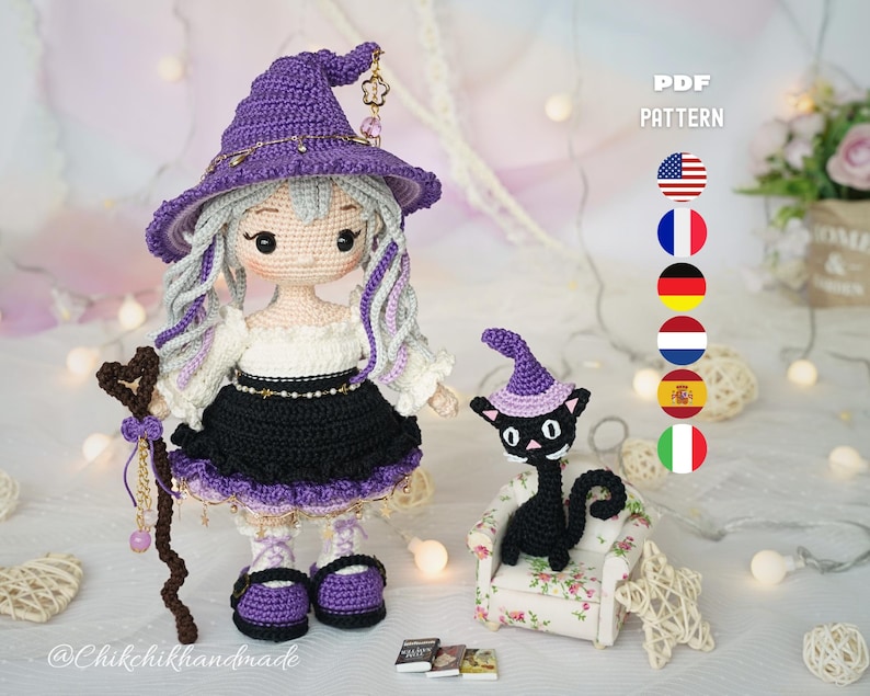 May include: A crochet doll with light skin, long wavy gray hair, and a purple witch hat, wearing a white blouse and black skirt. The doll holds a brown staff with a heart top. Beside it sits a small black cat in a purple hat on a floral chair.