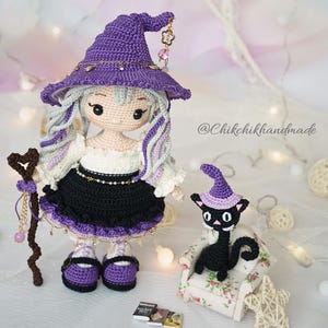 May include: A handcrafted crochet doll with long light gray hair wearing a purple witch hat, a white blouse, and a black skirt. The doll holds a brown heart-topped staff. Beside it sits a small black crochet cat wearing a purple hat on a chair.