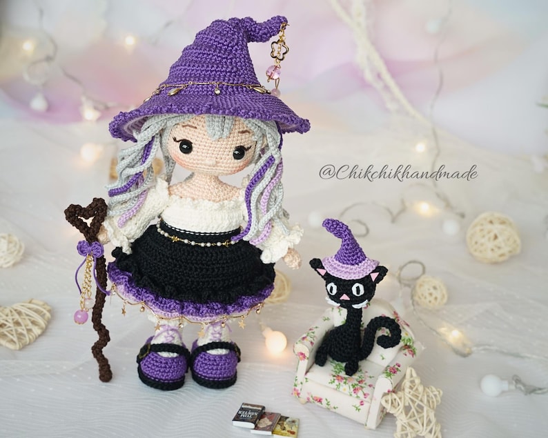 May include: A handcrafted crochet doll with long light gray hair wearing a purple witch hat, a white blouse, and a black skirt. The doll holds a brown heart-topped staff. Beside it sits a small black crochet cat wearing a purple hat on a chair.