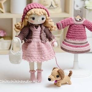 May include: A crocheted doll wearing a pink dress, brown cardigan, and a pink hat. The doll is holding a white crocheted bag and a leash attached to a brown crocheted dachshund. The doll is standing on a white surface. The image also includes a pink crocheted dress on a white stand.