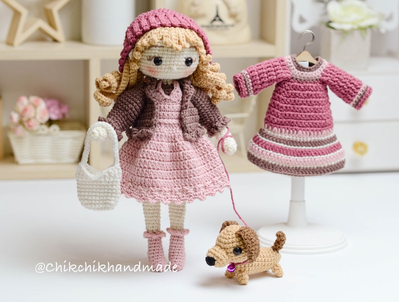 May include: A crocheted doll wearing a pink dress, brown cardigan, and a pink hat. The doll is holding a white crocheted bag and a leash attached to a brown crocheted dachshund. The doll is standing on a white surface. The image also includes a pink crocheted dress on a white stand.