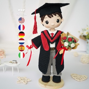 Puede incluir: Una muñeca de ganchillo vestida con una toga de graduación negra con estola roja, un birrete negro con borla roja y pantalones grises. La muñeca sostiene un diploma blanco atado con una cinta roja y un ramo de flores de ganchillo rojas y blancas.