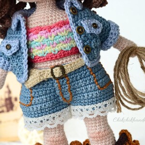 K&ouml;nnte beinhalten: Eine geh&auml;kelte Puppe, die eine Jeansjacke, ein regenbogenfarbenes Oberteil und Jeans-Shorts mit wei&szlig;em Spitzenbesatz tr&auml;gt. Die Puppe h&auml;lt ein braunes Seil.
