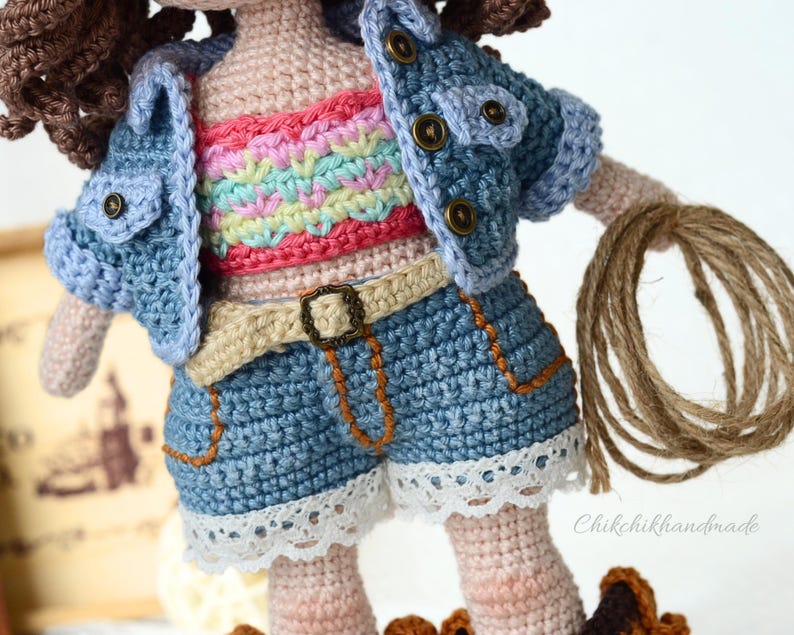 K&ouml;nnte beinhalten: Eine geh&auml;kelte Puppe, die eine Jeansjacke, ein regenbogenfarbenes Oberteil und Jeans-Shorts mit wei&szlig;em Spitzenbesatz tr&auml;gt. Die Puppe h&auml;lt ein braunes Seil.