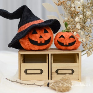 May include: Two crocheted pumpkins, one wearing a black witch hat, sitting on a wooden shelf with two drawers. The pumpkins have smiling faces and are decorated with orange and black yarn.