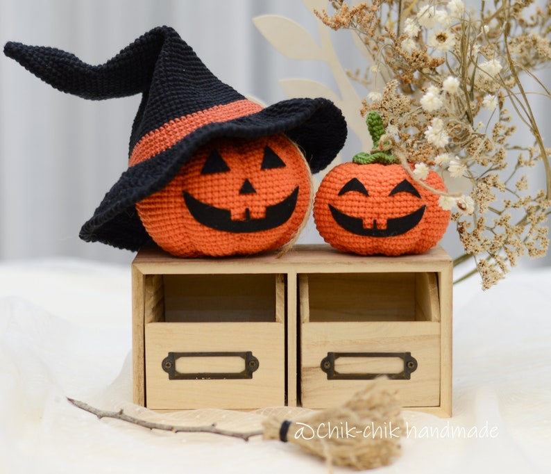 May include: Two crocheted pumpkins, one wearing a black witch hat, sitting on a wooden shelf with two drawers. The pumpkins have smiling faces and are decorated with orange and black yarn.