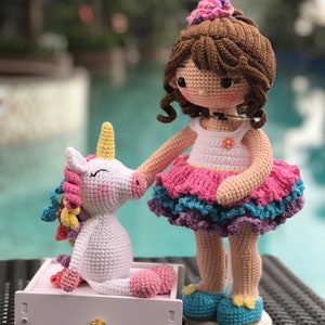 May include: A crocheted doll with brown hair and a pink and blue dress stands next to a white crocheted unicorn with a rainbow mane. The doll is wearing blue shoes and has a flower in her hair. The unicorn is sitting on a white drawer with a gold knob. The text "CHIK-CHIK HANDMADE" is visible on the bottom right corner of the image.